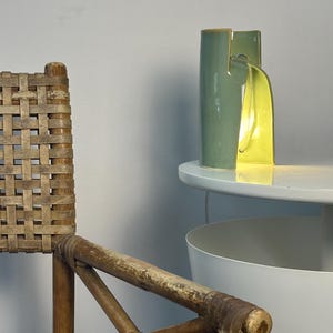May include: A cylindrical ceramic table lamp with a sage green exterior and a bright yellow interior. The lamp features a vertical slit design that allows light to emit from the center, casting a warm glow onto the surface it rests upon.