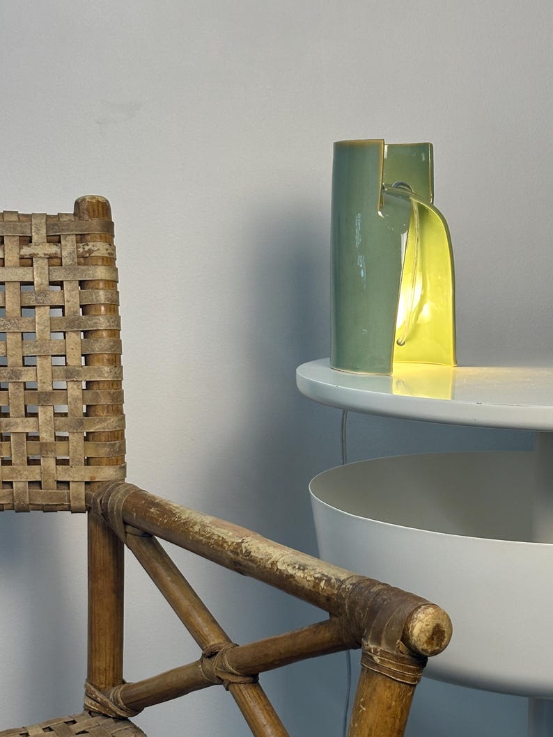 May include: A cylindrical ceramic table lamp with a sage green exterior and a bright yellow interior. The lamp features a vertical slit design that allows light to emit from the center, casting a warm glow onto the surface it rests upon.