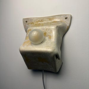 May include: A wall-mounted light fixture featuring a sculptural, off-white ceramic base with a mottled, textured glaze in shades of cream and yellow. A single, spherical, frosted glass bulb is centered on the front, with a thin power cord extending downward.
