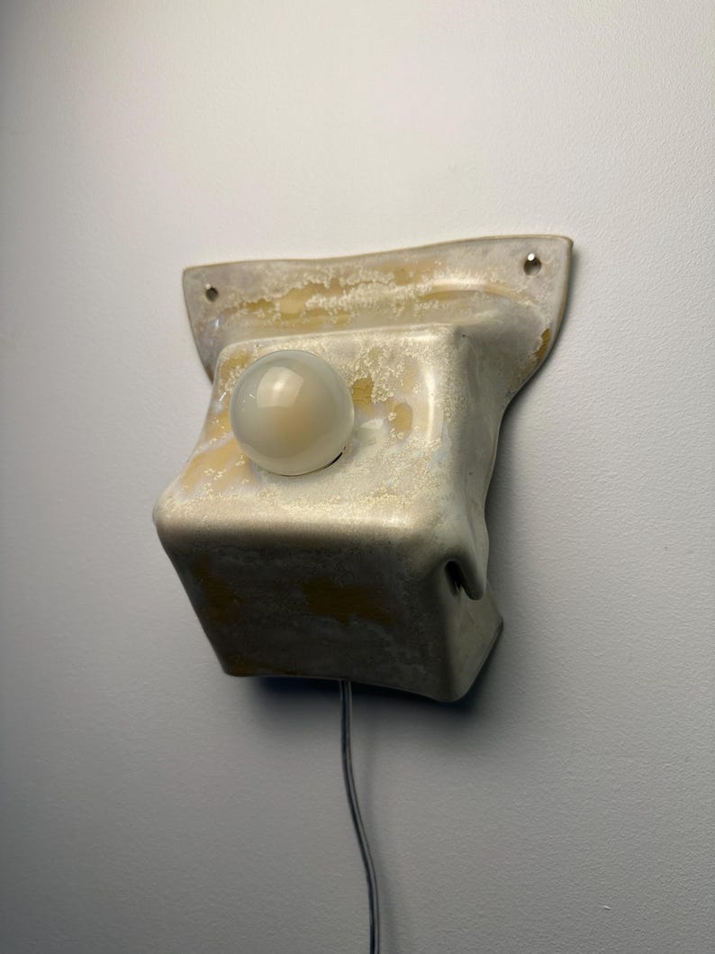 May include: A wall-mounted light fixture featuring a sculptural, off-white ceramic base with a mottled, textured glaze in shades of cream and yellow. A single, spherical, frosted glass bulb is centered on the front, with a thin power cord extending downward.