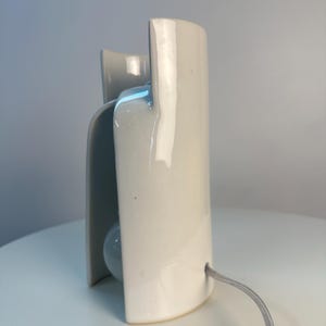 May include: A glossy white ceramic table lamp with a curved, sculptural silhouette. The lamp features a smooth, reflective surface and a light gray power cord extending from the side. The design consists of overlapping vertical panels.