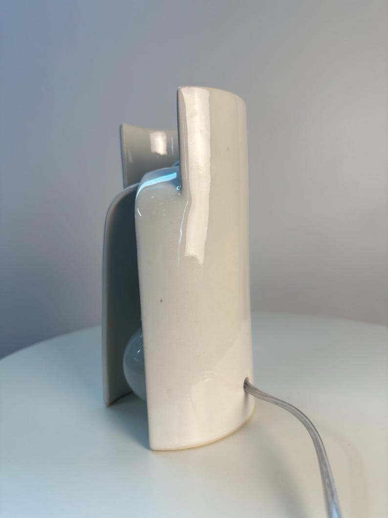 May include: A glossy white ceramic table lamp with a curved, sculptural silhouette. The lamp features a smooth, reflective surface and a light gray power cord extending from the side. The design consists of overlapping vertical panels.