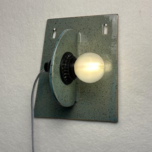 May include: A wall-mounted light fixture featuring a speckled light blue ceramic backplate with two rectangular cutouts. A perpendicular ceramic disc holds a black socket with a glowing round light bulb. A thin gray power cord extends from the side.