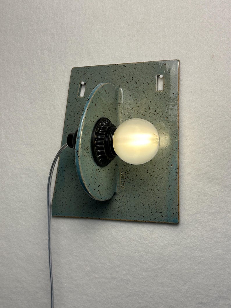 May include: A wall-mounted light fixture featuring a speckled light blue ceramic backplate with two rectangular cutouts. A perpendicular ceramic disc holds a black socket with a glowing round light bulb. A thin gray power cord extends from the side.