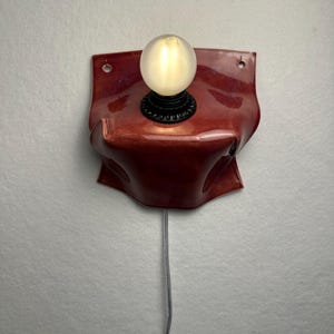 Puede incluir: Una l&aacute;mpara de pared con una base de cer&aacute;mica de color rojo oscuro brillante y forma org&aacute;nica plegada. El aplique incluye un casquillo negro con una bombilla redonda esmerilada que emite una luz c&aacute;lida, con un cable fino que cuelga hacia abajo.