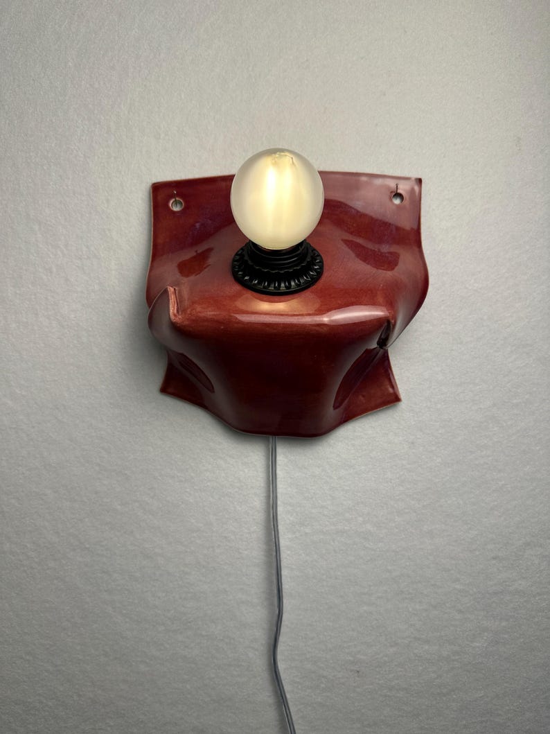 Puede incluir: Una l&aacute;mpara de pared con una base de cer&aacute;mica de color rojo oscuro brillante y forma org&aacute;nica plegada. El aplique incluye un casquillo negro con una bombilla redonda esmerilada que emite una luz c&aacute;lida, con un cable fino que cuelga hacia abajo.