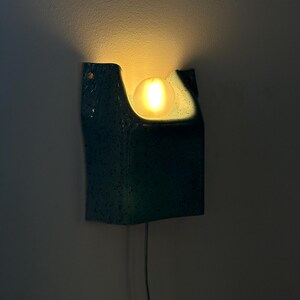 May include: A wall-mounted sconce featuring a teal, speckled ceramic housing with a curved, open top design. A single, glowing, spherical light bulb is positioned within the central cutout, casting a warm, soft light against the wall surface.