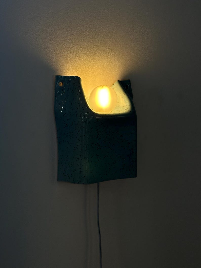 May include: A wall-mounted sconce featuring a teal, speckled ceramic housing with a curved, open top design. A single, glowing, spherical light bulb is positioned within the central cutout, casting a warm, soft light against the wall surface.