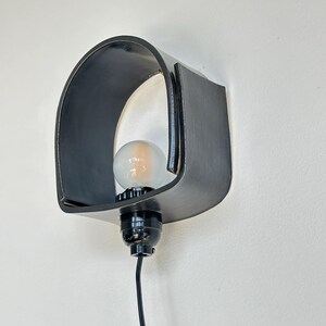 Puede incluir: Una l&aacute;mpara de pared negra con una pantalla curva en forma de arco. La l&aacute;mpara tiene una base negra y una bombilla visible. El dise&ntilde;o es minimalista y moderno, adecuado para la iluminaci&oacute;n interior.