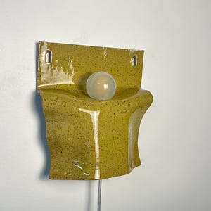 May include: A wall-mounted ceramic lamp featuring a glossy, mustard-yellow glaze with dark speckles. The fixture has a curved, folded form with two rectangular mounting holes at the top and a single, spherical, frosted white glass light bulb.