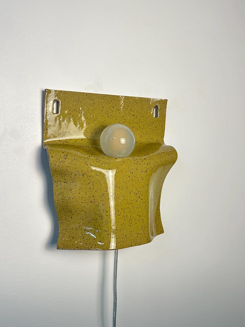 May include: A wall-mounted ceramic lamp featuring a glossy, mustard-yellow glaze with dark speckles. The fixture has a curved, folded form with two rectangular mounting holes at the top and a single, spherical, frosted white glass light bulb.