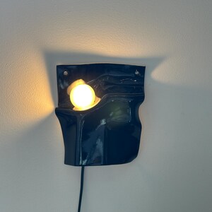 Puede incluir: Un aplique de pared de cer&aacute;mica azul oscuro con forma ondulada, acabado brillante y dos orificios de montaje en la parte superior. Una bombilla esf&eacute;rica de luz c&aacute;lida se aloja en la forma org&aacute;nica curva, incluyendo un cable de alimentaci&oacute;n negro.