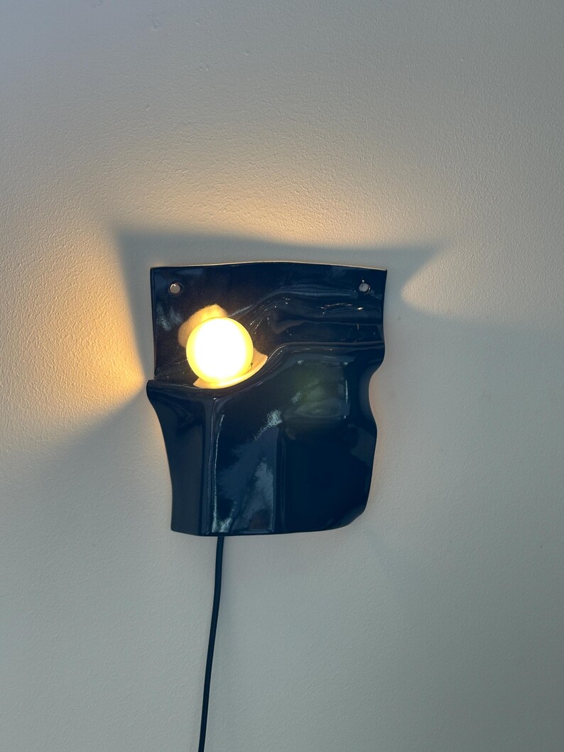 Puede incluir: Un aplique de pared de cer&aacute;mica azul oscuro con forma ondulada, acabado brillante y dos orificios de montaje en la parte superior. Una bombilla esf&eacute;rica de luz c&aacute;lida se aloja en la forma org&aacute;nica curva, incluyendo un cable de alimentaci&oacute;n negro.