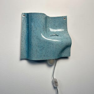 May include: A light blue wall sconce with a speckled finish and a wavy, sculptural form. The ceramic fixture features two mounting holes at the top corners, a central metal loop, and a downward-facing light bulb with a clear cord and switch.