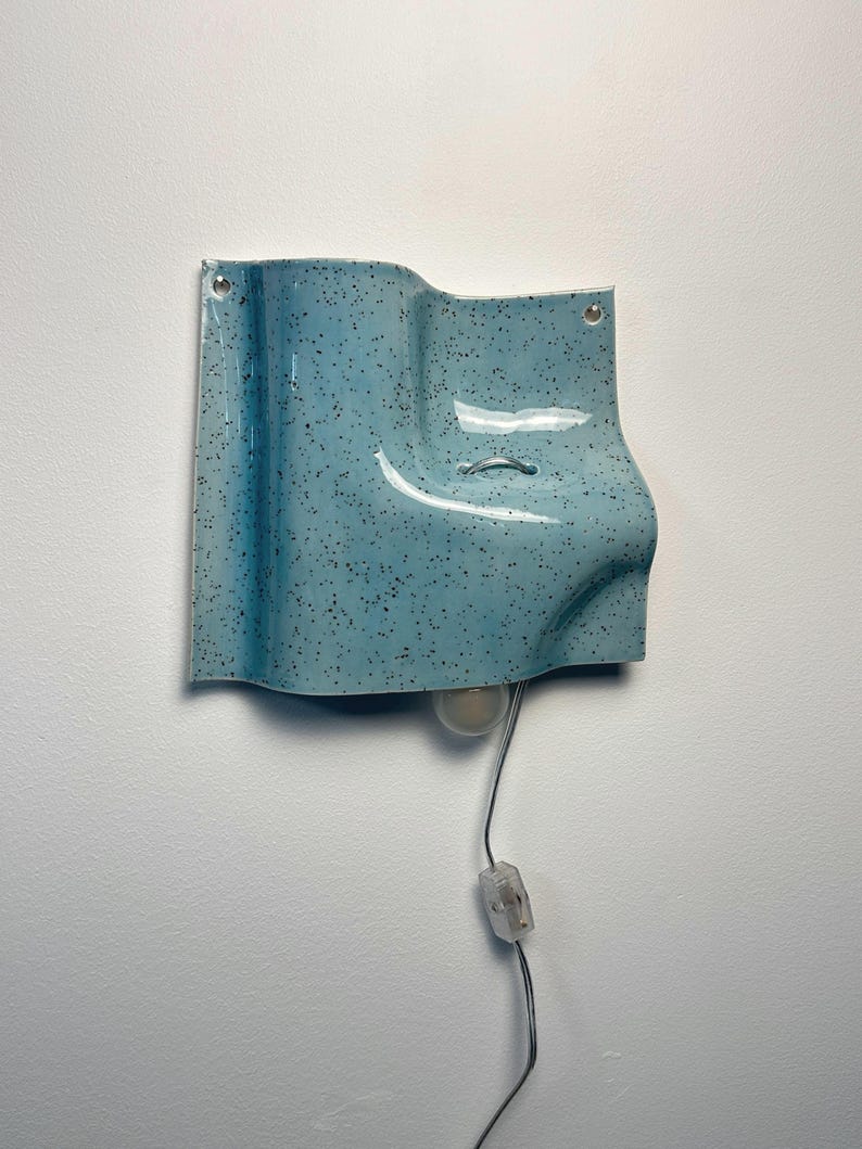 May include: A light blue wall sconce with a speckled finish and a wavy, sculptural form. The ceramic fixture features two mounting holes at the top corners, a central metal loop, and a downward-facing light bulb with a clear cord and switch.