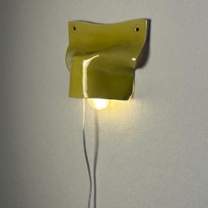 May include: A yellow wall sconce with a unique, folded design. The light fixture has a single bulb and a silver wire. The sconce is mounted on a light gray wall.