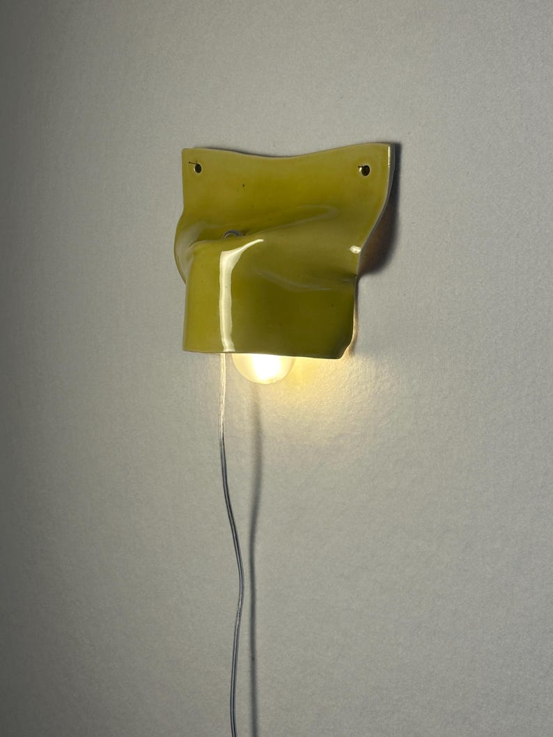 May include: A yellow wall sconce with a unique, folded design. The light fixture has a single bulb and a silver wire. The sconce is mounted on a light gray wall.