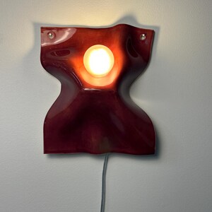 Puede incluir: Un aplique de pared con una pantalla de cer&aacute;mica de color rojo intenso, brillante y ondulada, que presenta una abertura circular central. Una bombilla encendida se sit&uacute;a en el centro y un cable el&eacute;ctrico gris sale de la parte inferior.