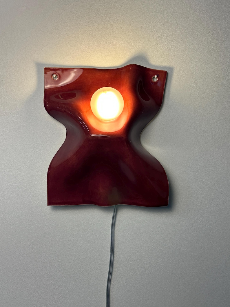 Puede incluir: Un aplique de pared con una pantalla de cer&aacute;mica de color rojo intenso, brillante y ondulada, que presenta una abertura circular central. Una bombilla encendida se sit&uacute;a en el centro y un cable el&eacute;ctrico gris sale de la parte inferior.