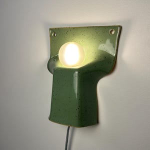 May include: A wall-mounted ceramic sconce in a speckled sage green glaze with a smooth, glossy finish. The fixture features a wide, flat top section with two mounting holes and a central, protruding curved base holding a glowing white orb bulb.