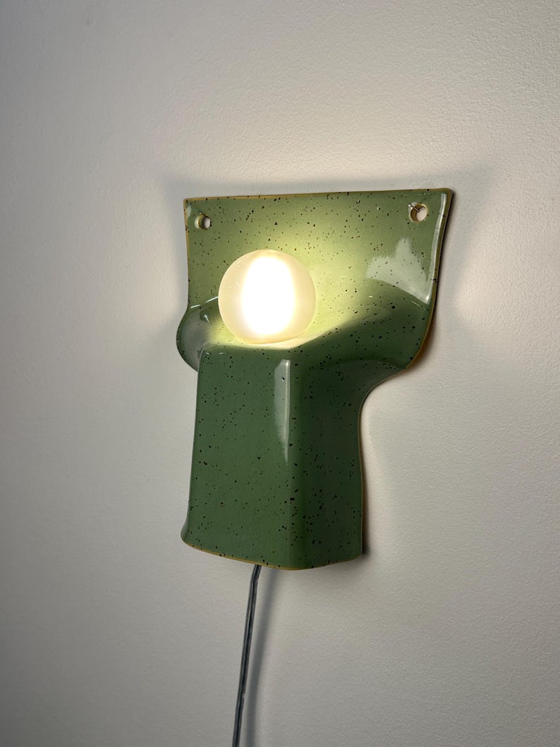 May include: A wall-mounted ceramic sconce in a speckled sage green glaze with a smooth, glossy finish. The fixture features a wide, flat top section with two mounting holes and a central, protruding curved base holding a glowing white orb bulb.