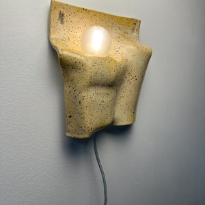 May include: A wall-mounted ceramic lamp with a speckled cream and brown finish. The fixture features a sculpted, torso-like form with a central circular opening holding a glowing, spherical white light bulb. A thin power cord hangs from the bottom.