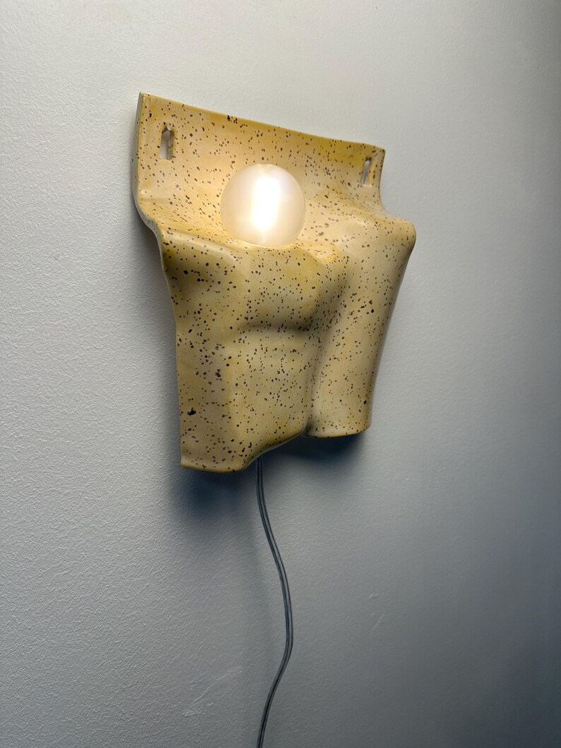 May include: A wall-mounted ceramic lamp with a speckled cream and brown finish. The fixture features a sculpted, torso-like form with a central circular opening holding a glowing, spherical white light bulb. A thin power cord hangs from the bottom.