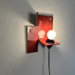 Pode incluir: Uma arandela de parede moderna com acabamento vermelho brilhante. A lumin&aacute;ria apresenta um design retangular com uma prateleira que suporta uma l&acirc;mpada redonda. Um cabo preto se estende para baixo. A arandela &eacute; montada em uma parede cinza.