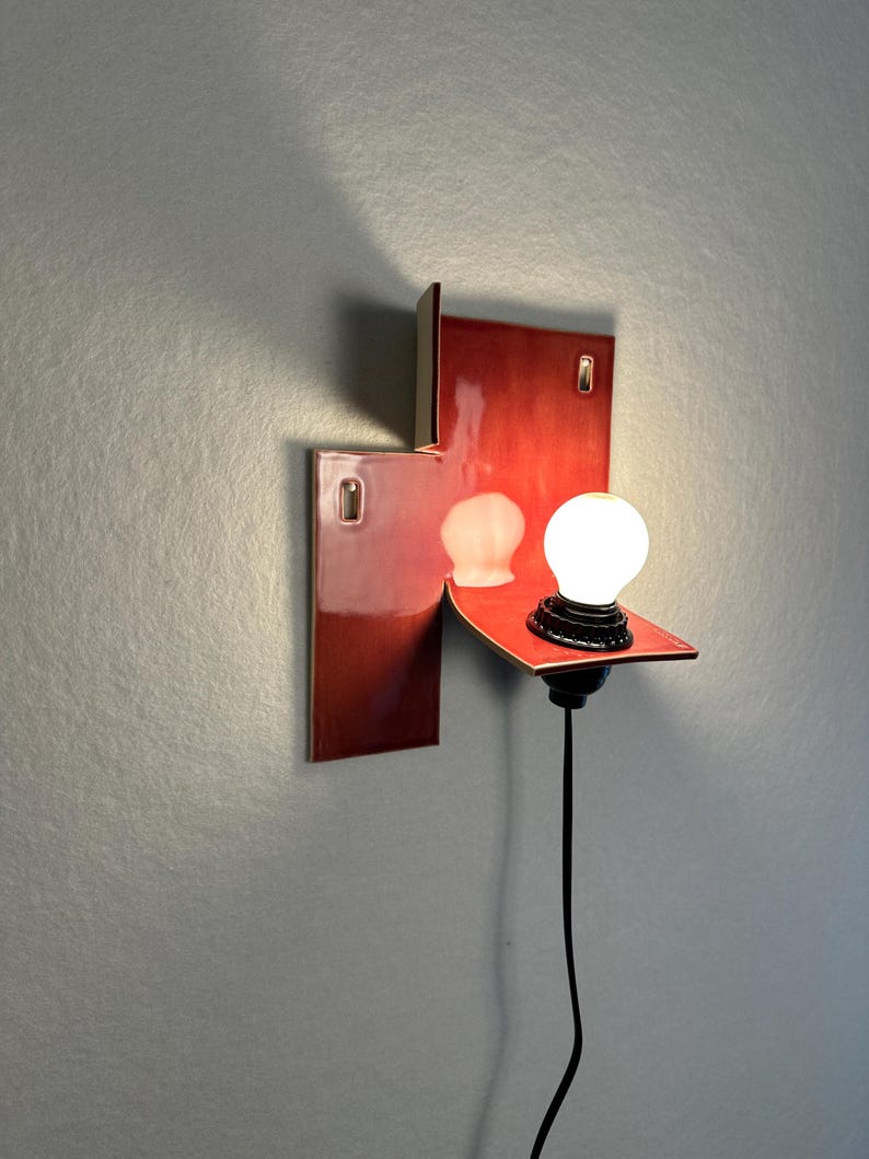 Pode incluir: Uma arandela de parede moderna com acabamento vermelho brilhante. A lumin&aacute;ria apresenta um design retangular com uma prateleira que suporta uma l&acirc;mpada redonda. Um cabo preto se estende para baixo. A arandela &eacute; montada em uma parede cinza.
