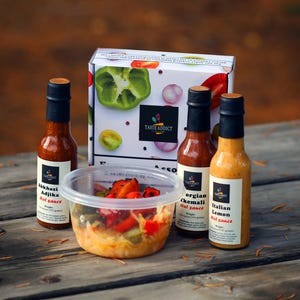 May include: A collection of hot sauces and a container of food on a wooden surface. The sauces are in glass bottles with black caps and labels. The box has a green pepper and the words "TASTE ADDICT". The food container is clear and filled with vegetables.