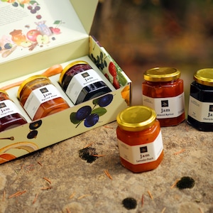 May include: A gift box filled with jars of jam, featuring flavors like spiced pear, plum, and cherry amarena. The jars have gold lids and white labels. The box is decorated with fruit illustrations.