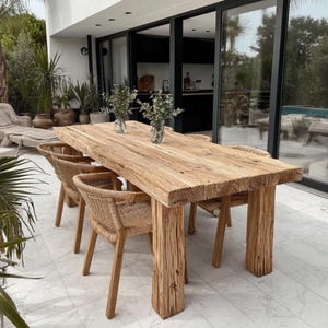 Puede incluir: Una mesa de comedor rectangular de madera rústica con borde natural, sostenida por gruesas patas cuadradas. Cuatro sillas de mimbre tejido están dispuestas alrededor de la mesa. Dos pequeños jarrones de cristal con vegetación sobre la mesa. La mesa mide unos 244 cm de largo.
