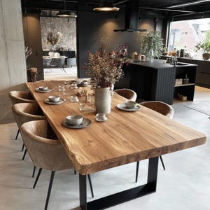 Könnte beinhalten: Ein großer, rechteckiger Esstisch aus Holz mit natürlicher Kante, gedeckt für eine Mahzeit. Der Tisch wird von schwarzen Metallbeinen getragen und ist von gepolsterten Stühlen umgeben. Eine Vase mit Trockenblumen steht in der Mitte, mit Gedecken darum.
