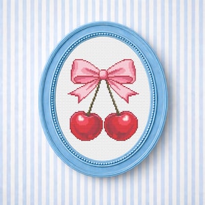 May include: Cross-stitch image of two red cherries with green stalks and a pink bow, presented in a light blue oval frame. The background features a white canvas with blue and white stripes.