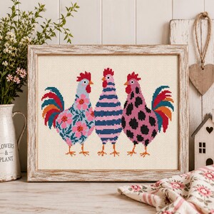 Puede incluir: Un bordado en punto de cruz enmarcado que muestra tres gallos de pie uno al lado del otro. El gallo de la izquierda tiene un cuerpo azul con flores rosas, el central tiene rayas horizontales azules y rosas, y el de la derecha es rosa con manchas negras.