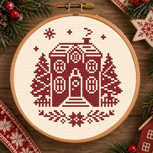 Può includere: Un telaio da ricamo circolare in legno contenente un motivo a punto croce su tessuto color crema. Il design presenta una casa rosso scuro con camino, circondata da due alberi di pino e piccoli motivi a fiocco di neve, in stile pixel.