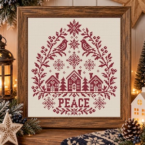 May include: A framed cross-stitch embroidery featuring a dark red design on a cream fabric background. The pattern displays three houses, two birds, snowflakes, and foliage arranged in a teardrop shape with the word PEACE at the bottom.