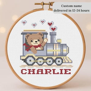 May include: Cross-stitch embroidery in a wooden hoop depicts a teddy bear driving a train, with hearts above. The name "CHARLIE" is stitched in red below the train. The text "Custom name" is visible.