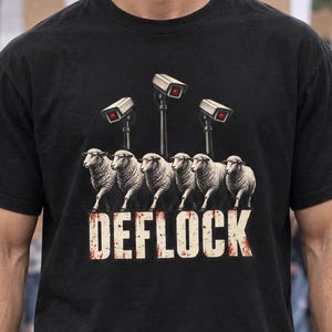 Könnte beinhalten: Ein schwarzes T-Shirt mit kurzen Ärmeln und einer Grafik von sechs weißen Schafen, die in einer Reihe unter drei Überwachungskameras laufen. Unter den Schafen steht das Wort DEFLOCK in einer abgenutzten weißen Schrift mit rötlicher Textur.