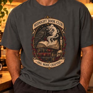 Può includere: Una t-shirt a maniche corte grigio antracite con una stampa grafica raffigurante una pila di libri, un corvo, un teschio, una candela e una figura spettrale. Il testo MIDNIGHT BOOK CLUB e ONE MORE CHAPTER è scritto su un nastro decorativo.