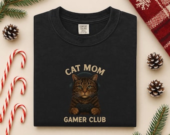 Puede incluir: Sudadera negra con el texto "CAT MOM GAMER CLUB" y un gráfico de un gato con auriculares y un mando de videojuegos. La sudadera está rodeada de adornos navideños.