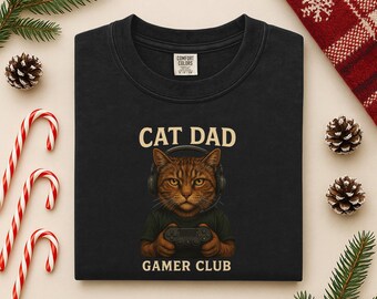 May include: Black crew neck sweatshirt with a graphic of a cat wearing headphones and holding a game controller. The text "CAT DAD" is above the cat, and "GAMER CLUB" is below.