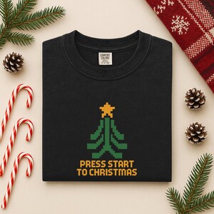 May include: Black crew neck sweatshirt featuring a green and yellow pixelated Christmas tree and the text "PRESS START TO CHRISTMAS". The sweatshirt is surrounded by festive decorations, including candy canes and pine cones.