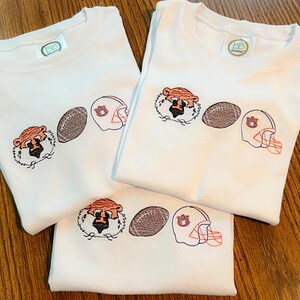 May include: Three white crew neck shirts with embroidered designs. Each shirt features a tiger head, a football, and a football helmet. The tiger head is orange and black, the football is brown, and the helmet is orange and blue.