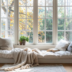 May include: A cozy window seat with a cream-colored cushion, pillows, and a knitted throw blanket. A small potted plant sits on the windowsill. The window offers a view of trees with yellow and green leaves. A bookshelf is on the right.