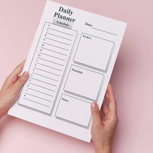 Könnte beinhalten: Ein weißes Daily Planner-Blatt mit Abschnitten für Zeitplan, Aufgaben, Prioritäten und Notizen. Der Planer wird vor einem rosa Hintergrund gehalten. Der Text "Daily Planner" steht oben.