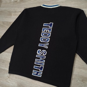 Peut inclure: Pull à manches longues noir avec le texte "TEDDY SMITH" en bleu et blanc dans le dos. Le pull a un col rayé blanc et bleu.