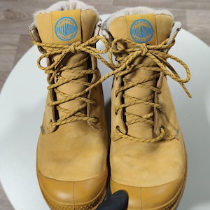 May include: Tan Palladium boots with a rubber toe cap and yellow laces. The boots feature a blue and white logo patch and a faux fur lining. These boots are designed for cold weather. The boots are approximately 20 cm in height.