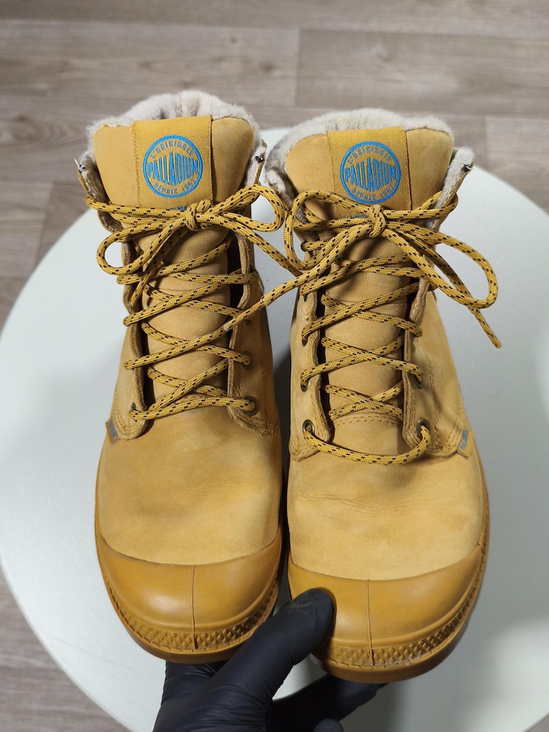 May include: Tan Palladium boots with a rubber toe cap and yellow laces. The boots feature a blue and white logo patch and a faux fur lining. These boots are designed for cold weather. The boots are approximately 20 cm in height.