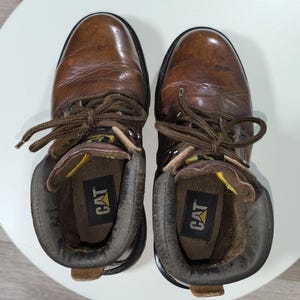 May include: A pair of brown leather work boots with laces, viewed from above. The boots have a worn appearance, with the brand name "CAT" visible inside. The boots are placed on a white surface.